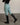 A model from the back wearing pro breeches in beachy green equestrian riding breeches with a black belt, paired with a black long-sleeve top and black riding boots, standing on a concrete floor against a neutral beige wall.