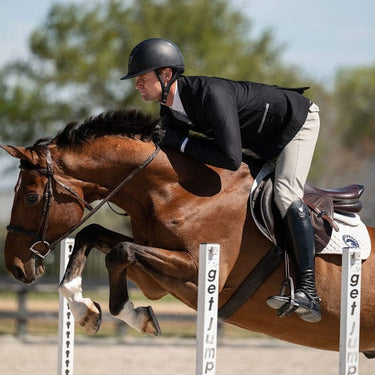 A rider in black equestrian attire and a black helmet sits on a brown horse jumping over a white jump obstacle in an outdoor equestrian arena.