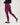 A model wearing high-waisted, form-fitting leggings in a raspberry color, paired with a black long-sleeve top and black slip-on sneakers, shown from a side-back angle against a plain white studio background.